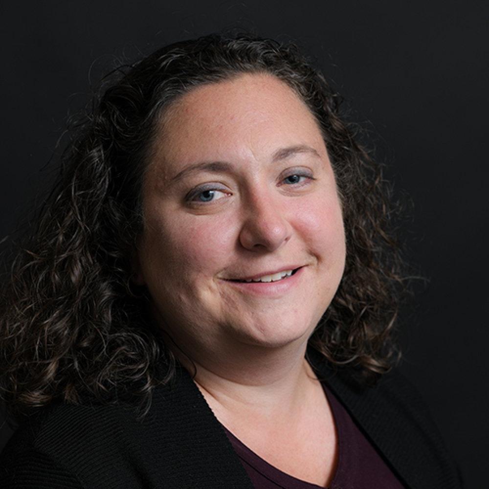 Kerry Paumi headshot, lady with dark curly hair wearing black top with black background